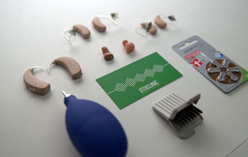 Modelos de aparelhos auditivos retroauriculares e intracanais sobre a mesa, com cartão da Otoclinic, pilhas e acessórios de limpeza